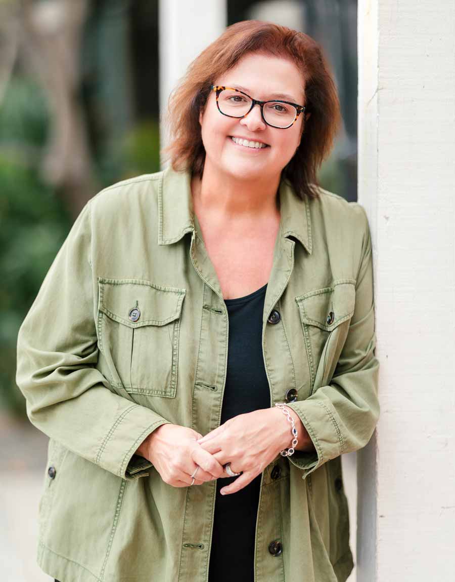 Portrait of HelenKay in a green button down shirt and glasses leaning against a white pillar